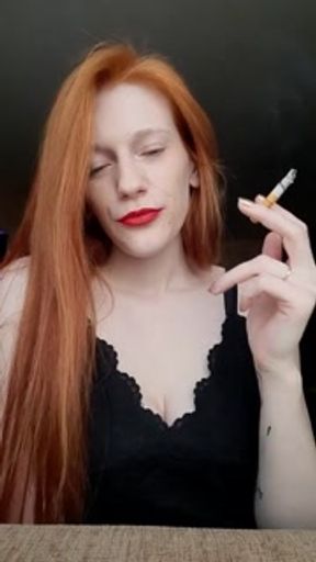 A girl with long red hair in a black dress and red lipstick smokes a cigarette with a brown filter