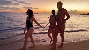Tropical Sunset Chastity Stroll - Reese the Ram is in full chastity and a latex hood, BondageFan70 is in his rigid metal exoskeleton and rubber hood. Their keeper, Delphine Mason, walks them along a beach.